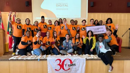 Group of women posing for photo in an auditorium stage. Part of the group is standing in the back, the other is is sitting in front. Most are wearing orange T-shirt. A flag saying Themis 30 is also seen at the bottom of the stage.