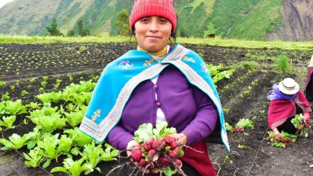Woman standing in a plantation holding vegetables. Woman is wearing a red hat, purple cardigan, and blue scarf on top.