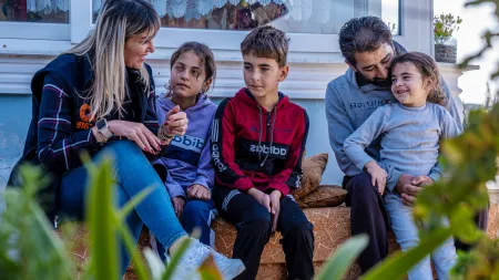 A blonde woman, three children, and a man sitting on the porch a house.