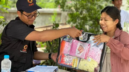 Man wearing hat dark blue hat and shirt with CARE's logo handing transparent plastic bag with baby items to woman. Woman is wearing light colored clothes, has dark straight hair, and is smiling.