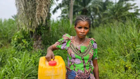 Woman wearing patterned dress, carrying a a water can, while standing in a green area.