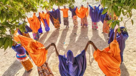 Group of women in a circle holding heads up. Women are wearing orange and blue veils and are standing in an outdoor area, under a tree.
