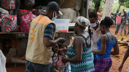 Man wearing jacket with CARE's logo, hat and face mask, handing a bag to woman wearing patterned dress in the context of a wider aid distribution.