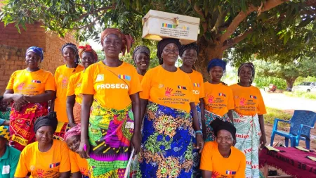 Women wearing orange t-shits with logos and patterned cloths at the bottom. Woman in the centre has a locked box balancing over her head.