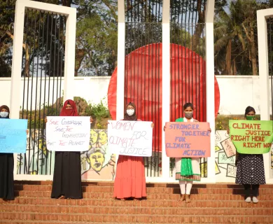 A group of Bangladeshi women stand in a line on a set of stairs. Each of them is wearing a face mask and holding a handmade sign with messages advocating for climate justice.
