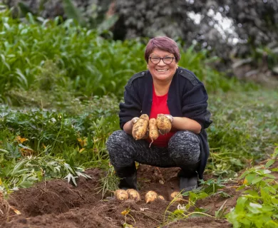 A short-haired Peruvian woman squats in a field and proudly holds up a few potatoes.