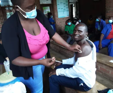 A man is sitting in a chair with his shirt pulled down to reveal his shoulder. A woman wearing a blue surgical face mask is wiping his upper arm with a cotton ball and preparing to give him a shot.