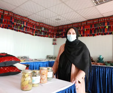 A woman wearing a head scarf and surgical face mask stands at a table with jarred and canned goods.