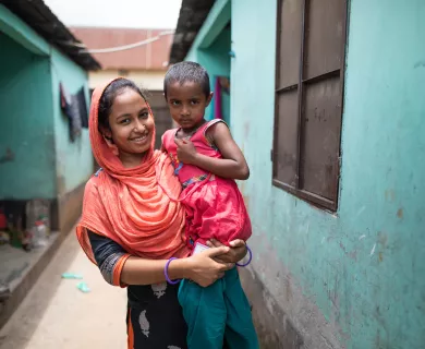 A woman smiles while holding a child in her arms. They are standing in an alley way with cheerful teal blue buildings on either side.