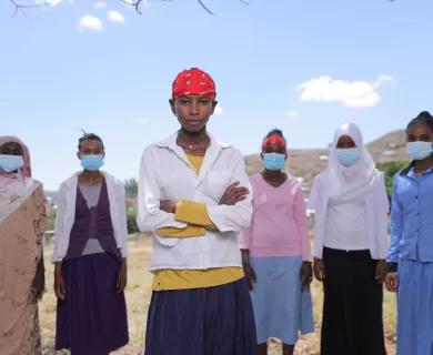 An Ethiopian girl wearing a red bandanna crosses her arms. Behind her are a line of women, all wearing blue surgical facemasks.