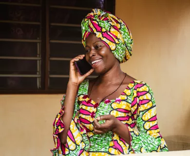 A woman wearing a brightly colored pink, yellow, and green dress and matching head wrap smiles cheerfully while she talks on a black mobile phone.