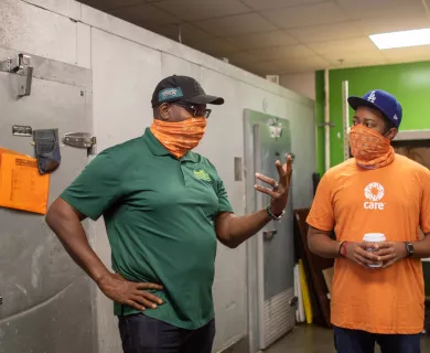 Two men, each wearing orange CARE face bandanas, talk while standing next to large walk-in food freezers.