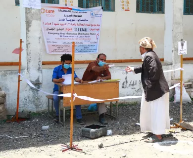 Two people sit behind a desk, each wearing a blue surgical mask. One of them is reaching to hand a woman a paper document.