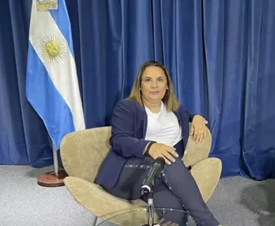 Woman looking serious to the camera, with blonde straight hair, sitting on a chair with the Argentinian flag in the background