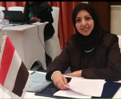 Woman smiling to the camera wearing black headscarf and clothes. Sitting at a table holding papers