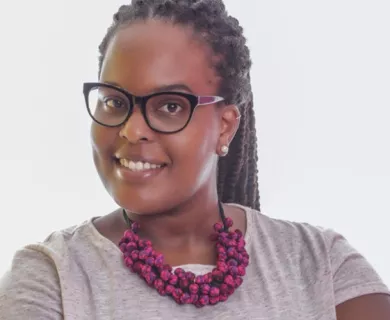 Headshot of woman smiling to the camera, wearing glasses and braided hair a in pony tail. Wearing a purple necklace and a grey shirt.