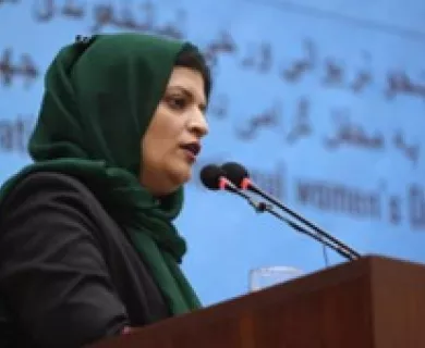 Woman speaking on microphone at stand. Wearing green headscarf and black shirt.
