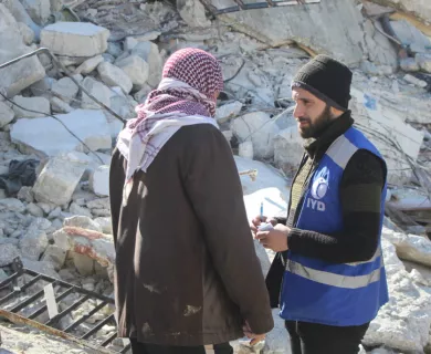 Syria_Man wearing IYD vest talking to Syrian man with red and white scarf in front of rubble