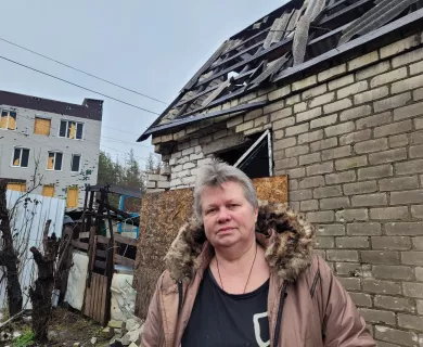 Woman with grey hair and pink coat in front of house with destroyed roof