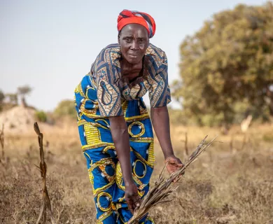 A woman wearing blue and yellow patterned clothes shows her withered crops in the field. Dried grass and tress in the background.