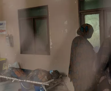 A person lying on a cot, covered with a patterned blanket, while two women in patterned shawls stand nearby in a dimly lit room with CARE and SHF signage.