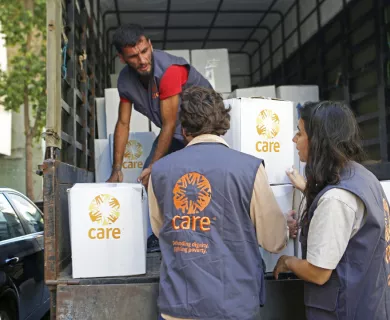 CARE aid workers load boxes marked with CARE logo onto a truck. Three people visible, wearing CARE vests. Scene shows humanitarian supply distribution in progress.
