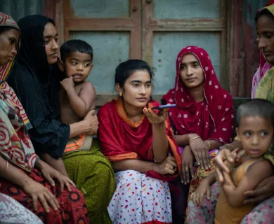Group of South Asian women and children sitting together. Women wear colorful headscarves. Young woman in red holds smartphone. 