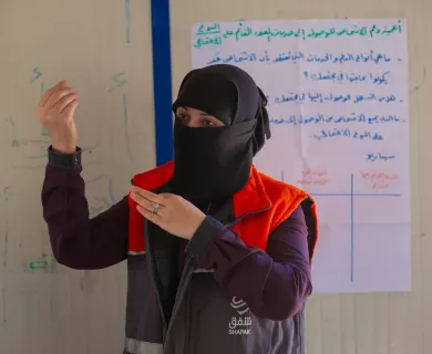 Woman wearing black head scarf and face cover with gesticulating in a room with large information paper up on the wall.