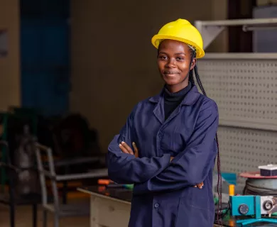 Young woman wearing dark blue work jumpsuit and yellow construction head, with arms crossed and gentle smile to the camera