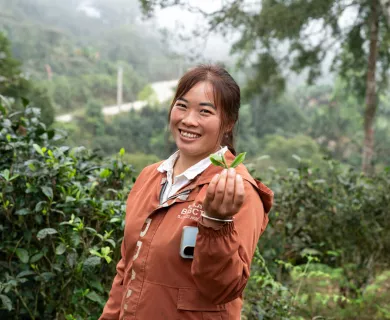 Woman wearing orange jacket, holding a tea leaf and smiling at the camera, while standing in a green area