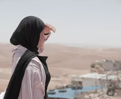 Side view of woman wearing black head scarf and light pink shirt, standing outdoors, covering her eyes from the sun with left hand